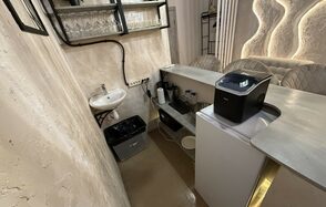 Лофт SUNRISE(Восход Солнца). Просторное помещение на 15 гостей. Маленькая кухня с барной стойкой, бежевые стены, белая раковина, металлические полки для бокалов, встроенный освежитель, мусорное ведро, белая техника, настенное освещение с изогнутыми линиями.