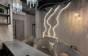 Лофт SUNRISE(Восход Солнца). Просторное помещение на 15 гостей. Современное помещение с барной стойкой и мягкими креслами, декоративное освещение с подвесными лампами, акцентная стена с текстурой и подсветкой, открытые полки с растениями, светло-серые и бежевые оттенки, минималистичный интерьер.