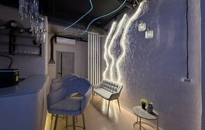 Лофт SUNRISE(Восход Солнца). Просторное помещение на 15 гостей. Небольшое помещение с декоративной стеной и неоновым освещением. Барная стойка с барными стульями. Мягкие кресла и круглый столик с растением. Подвесные светильники. Потолок с декоративной проводкой и вентиляцией. Стены текстурированные.