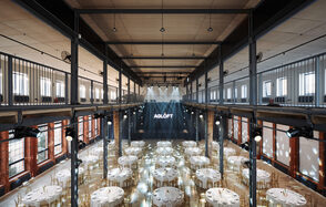AG LOFT. Grand Hall (АГ Лофт. Большой зал) . Большое открытое пространство с высоким потолком и металлическими балками, круглыми столами с белыми скатертями и стульями. Внизу сцена с подсветкой и мониторами. Многоуровневое расположение с балконом. Стены из красного кирпича с большими окнами. Надпись "AGLOFT".