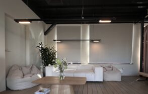 Пространство с террасой до 50 человек. Современная гостиная с деревянным полом, белым диваном, бежевыми креслами-мешками, деревянным журнальным столиком. Белые жалюзи на окнах, две подвесные лампы сверху. Черные потолочные балки, зеленое растение в углу, на столе ваза с цветами.