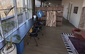 Пространство с террасой до 50 человек. Пространство в стиле лофт с большими окнами, вдоль которых стоит длинный стол с черными офисными стульями. На полу деревянный настил, коврик с черно-белыми полосами и красными элементами, разделение из деревянных панелей. Декоративные растения.