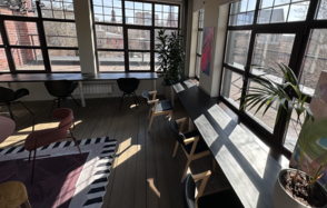Пространство с террасой до 50 человек. Просторное помещение с большими окнами, светлое дерево на полу, черные и розовые кресла, длинные столы вдоль окон, несколько растений, абстрактная картина на стене, ковёр с геометрическим узором, кирпичная стена у окна.