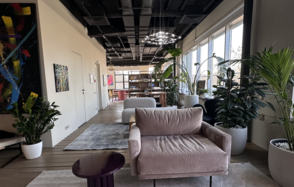 Пространство с террасой до 50 человек. Современное пространство для мероприятий с большим окном, серым диваном, фиолетовым столиком, множеством зелёных растений в кашпо. Потолок тёмный с видимыми вентиляционными системами, освещение в виде дизайнерских светильников, стены бежевые с художественными картинами.