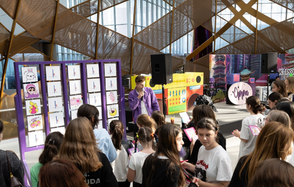 Кунцево Плаза. Современное пространство для мероприятий с высокими потолками и стеклянными стенами. Панель с рисунками и ведущей, выступающей у микрофона. Множество людей, в основном подростки. Цветная декоративная отделка и панели с изображениями.