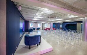 АКИ.лаб пространство для мероприятий. Просторное помещение с серым бетонным полом и подвесным потолком. Стулья из прозрачного пластика расположены перед подиумом с двумя синими креслами. Стены темные с большими экранами. Освещение встроенное, равномерное.