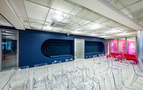 АКИ.лаб пространство для мероприятий. Просторное помещение с белым потолком и синей стеной, украшенной геометрическими панелями. Прозрачные пластиковые стулья расположены рядами. Справа стеклянная стена с розовой подсветкой. Пол светлого оттенка, потолок с встраиваемыми светильниками.