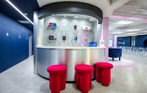 АКИ.лаб пространство для мероприятий. Современное помещение с белым столом полукруглой формы и тремя ярко-красными пуфами. Стены тёмно-синие с белыми дверями, пол серый. Подвесные светильники, стеклянные выставочные элементы на стене, прозрачные стулья, информационные таблички.