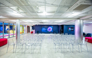 АКИ.лаб пространство для мероприятий. Просторное помещение с белым потолком и бетонным полом. Прозрачные пластиковые стулья расставлены рядами перед экраном на синей стене. Яркое электрическое освещение, стеклянные колонны, цветные стены сбоку, яркие текстильные диваны.