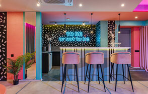 Лофт Шекспир. Стильный дизайнерский зал. Барная зона с высокими стульями и барной стойкой, подсвеченной неоновым текстом «TO BE or not to be». Стены с черно-белым узором, потолочное освещение, яркие цвета, элементы розового и голубого, зеркало, растения в углу.