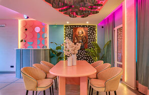 Лофт Шекспир. Стильный дизайнерский зал. Розовый овальный стол с бежевыми стульями, на стене портрет в рамке, барная стойка с круглыми элементами, розовый и голубой декор, растения, многоцветное освещение, шторы, потолочный элемент с волнениями.