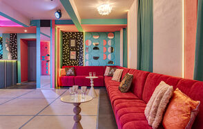 Лофт Шекспир. Стильный дизайнерский зал. Пространство с длинным красным диваном с подушками, круглый столик с бокалами, яркие стены с геометрическими узорами, различные картины, декоративное освещение, разноцветные стены и потолок, плиточный пол светлых оттенков.