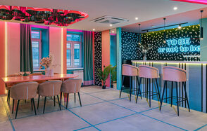 Лофт Шекспир. Стильный дизайнерский зал. Современное праздничное пространство с барной стойкой и столовой зоной. Бар с высокими стульями, неоновая надпись, обеденный стол с мягкими стульями. Яркое декоративное освещение, разноцветные стены, большие окна с шторами, растения в горшках.