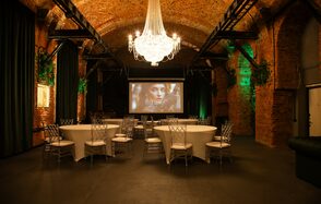 Arsenal Loft(Арсенал Лофт). Винтажное пространство с кирпичными сводчатыми потолками и стенами. Круглые столы с белыми скатертями, прозрачные стулья. Большая люстра и зелёная подсветка. Экран с проекцией. Тёмный пол. Живые растения на стенах.