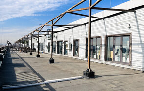 Зал под кулинарные мастер классы и мероприятия. Терраса под открытым небом с деревянным навесом, витражными окнами и кондиционером на белой стене здания. Деревянные балки поддерживают структуру, на полу бетон. Светлый день, частично облачное небо.