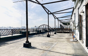 Зал под кулинарные мастер классы и мероприятия. Открытая терраса с бетонным полом, металлическими каркасами и перилами, деревянными столами и стульями справа. Видна часть здания с деревянной отделкой и кондиционером. Потолочные гирлянды на каркасах, дневное освещение.