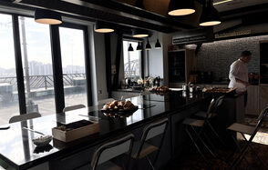 Зал под кулинарные мастер классы и мероприятия. Кухонное пространство с длинным островом из темного материала, окружённым складными стульями. На столе расположены блюда и столовые приборы. Подвесные светильники, большие окна с видом на город, светлые стены, зона приготовления с плитой и вытяжкой, текст на стене.