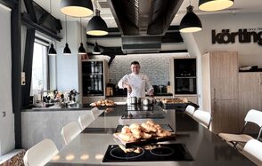 Зал под кулинарные мастер классы и мероприятия. Кухонное пространство с островом, варочная панель, современное освещение, подвесные светильники, барные стулья, деревянные шкафы, стеклянная дверь, выпечка на прилавке, человек в белой униформе, металлическая вытяжка, стена с кирпичным узором, логотип на стене.