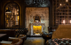 Каминный Зал Tangiers lounge Pokrovka(Танжерс Лаундж Покровка). Интерьерное пространство с темной атмосферой и кожаной мебелью. Центральный элемент — искусственный каменный камин с зеркалом, свечами и часами. Над камином — большой черный люстр. Показаны светильники с теплым освещением.