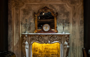 Каминный Зал Tangiers lounge Pokrovka(Танжерс Лаундж Покровка). Камин с резным каменным порталом, украшенным декоративным орнаментом. Над камином зеркало в золочёной раме, по бокам металлические канделябры. Внутри камина подсветка, стены с бежевой текстурой. На камине стоит старинные часы.