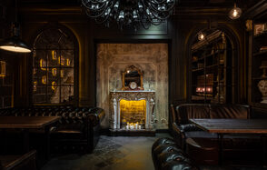 Каминный Зал Tangiers lounge Pokrovka(Танжерс Лаундж Покровка). Интерьер в классическом стиле с камином, кожаными диванами темного цвета и деревянными столами. Стены украшены книжными полками и статуей бюста. Потолочная люстра и несколько ламп накаливания создают теплое освещение.