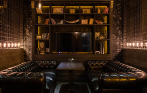 Каминный Зал Tangiers lounge Pokrovka(Танжерс Лаундж Покровка). Лофт-пространство с чёрными кожаными диванами, деревянным столом, стеллажом с книгами и декором, телевизором. Стены кирпичные, освещение тёплое, из лампочек над стеллажом и по бокам. Тёмная цветовая гамма, интерьер в классическом стиле.