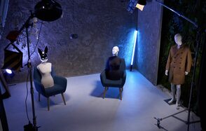 WhiteFox Studio(Студия Белая Лиса). Серое помещение с бетонными стенами, два манекена в креслах, один манекен в пальто стоит, стул с текстильной обивкой, неоновая лампа, потолочные и напольные светильники, фон из растений, светлая поверхность пола, студийное оборудование.