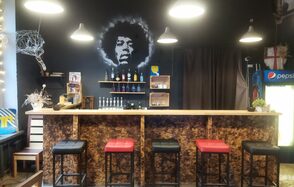 Лофт-Бар. Барная стойка с высокими стульями в черных и красных тонах, темная стена с изображением человека и полками с бутылками, подвесные светильники, холодильник с напитками, деревянные элементы декора, закрытая плотной темной шторой ниша.
