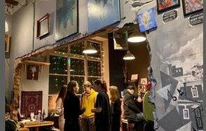 Лофт-Бар. Арт-пространство с обеденной зоной, стены украшены картинами, окно с гирляндами, столы и стулья из дерева, неоновые таблички, подвесные светильники, кирпичная кладка, разнообразные цветовые акценты, несколько человек общаются.