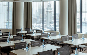 Конференц-зал Экспресс Махараджей. Конференц-зал с большими окнами, прямоугольные столы с расставленными бутылками воды и стаканами. Серые стулья на колесиках. На улице зимний пейзаж. Светлые шторы, нейтральные тона интерьера, хорошее естественное освещение.