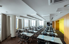 Конференц-зал Экспресс Махараджей. Конференц-зал с рядами столов, окружённых черными стульями. Ярко-желтая стена слева, окна с бежевыми шторами справа. Белый потолок с встроенным освещением. Темный ковролин на полу. Пластиковые бутылки с водой на столах.