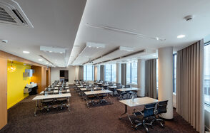 Конференц-зал Экспресс Махараджей. Конференц-зал с прямоугольной планировкой, большого размера окна. Ряд столов с черными стульями. Белые потолочные светильники, стены из светлого дерева с желтыми акцентами. Ковровое покрытие темного цвета. Шторы бежевые.