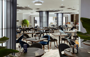 Итальянский ресторан Umberto(Умберто). Просторное помещение ресторана с расставленными квадратными столиками и темными стульями, большая барная стойка на заднем плане. Светлые стены и потолок с встраиваемыми светильниками. Декоративные растения, стеклянные и металлические элементы.