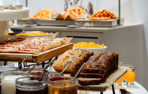 Итальянский ресторан Umberto(Умберто). Деревянные подносы с нарезанными пирогами на столе. Рядом банки с джемом и ёмкости для напитков. На заднем плане видны тарелки с пирожными и выпечкой. Пастельные оттенки, современное оформление, яркое освещение.