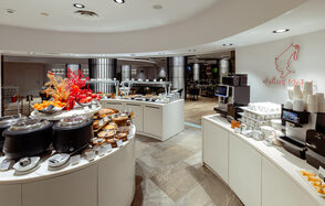 Итальянский ресторан Umberto(Умберто). Современная закусочная зона в помещении с круглой сервировкой. Белые стойки с выпечкой и суповыми станциями, аппарат для напитков. Открытая планировка, деревянное и плиточное покрытие. Яркое освещение, декоративные растения. Логотип на стене.