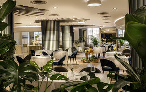 Итальянский ресторан Umberto(Умберто). Современное ресторанное пространство с круглыми столами, накрытыми белыми скатертями. Черные стулья, зелёные растения, мягкое освещение. Пол деревянный, колонны с декоративными полосами. Потолок с круглым освещением и вентиляцией.