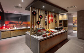 Ресторан Ibis Kitchen(Ибис Кухня). Ресторанное пространство с сервировочным островом. На столе блюда и напитки. Интерьер из дерева и камня. Настенные украшения: виниловые пластинки и постеры. Современное освещение. В тёплых тонах, доминируют коричневый и красный цвета.