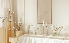 OLFI-Studio(ОЛФИ Студия). Фотография праздничного пространства с кремовыми стенами и текстильным декором на стене. На белоснежном столе в вазах стоят белые цветы и свечи. Справа и слева расположены круглые столики с вазами и цветами. Освещение мягкое, дневное.