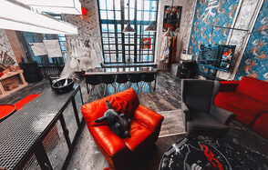 Лофт Делюкс. Лофт-пространство, декорированное в индустриальном стиле с большими окнами. В центре длинный деревянный стол с черными стульями. Красное и серое кресла, яркий диван. Декоративные элементы на стенах, большие подвесные светильники, темное напольное покрытие.