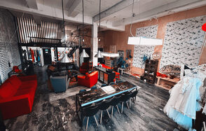 Лофт Делюкс. Лофт-пространство с бетонным потолком и колоннами, металлическими светильниками и деревянным полом. В центре длинный стол с черными стульями, кожаные кресла и красные диваны. На стенах декор и растения, повсюду яркое освещение.