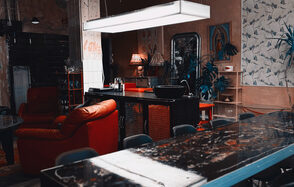 Лофт Делюкс. Лофт-пространство с барной стойкой, украшенной красными элементами. Красные мягкие кресла, светильники на стенах, под потолком прямоугольный светильник. Стена с растительным узором, деревянные и металлические детали интерьера, комнатные растения.