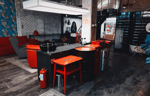 Лофт Делюкс. Лофт-пространство с барной стойкой из железных бочек и деревянным столом; кирпичные стены, деревянный пол, красная и серая мебель, большое центральное освещение; имеются диваны, кресла, раковина, огнетушитель; декорированные стены и стены с графическими рисунками.