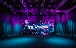 New Media (Нью Медиа) продакшн. Павильон с большим LED-экраном. Темное помещение с черными шторами и натяжным потолком с фиолетовой подсветкой. В центре светодиодный экран с цветным анимированным изображением и текстом "new media". Пол темного оттенка, система освещения закреплена на металлических конструкциях.
