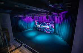 New Media (Нью Медиа) продакшн. Павильон с большим LED-экраном. Темное помещение с черным потолком и оборудованием для освещения. Стены оформлены черными шторами. На полу темное покрытие. Спереди большой экран с яркими графиками и текстом "newmedia". Освещение фиолетовое и голубое.