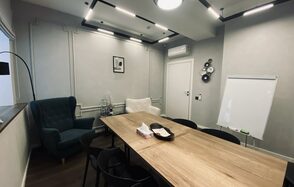 Дом Счастья. Современное офисное помещение с длинным деревянным столом и черными стульями. На стене висят декоративные элементы. Освещение потолочное, встроенное. В углу белая доска и кондиционер. Две мягкие кресла, бутылки с водой. Стены серые.