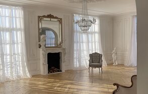 Арт-Плейс Фактура. Просторный лофт в классическом стиле. Просторное белое помещение с высоким потолком, украшенным люстрой. Паркеты из светлого дерева. Камин с декоративной рамой и скульптурами. Возле окна кресло серого цвета. Высокие окна занавешены легкими прозрачными шторами.