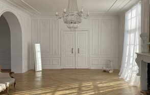 Арт-Плейс Фактура. Просторный лофт в классическом стиле. Светлая комната с паркетным полом, белыми стенами с декоративной лепниной и двойными дверями. Хрустальная люстра висит на потолке. Справа - камин и скульптура, слева - большое зеркало и арка. Под окном - занавески.