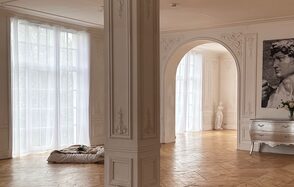 Арт-Плейс Фактура. Просторный лофт в классическом стиле. Просторное помещение с белыми резными стенами и колонной, паркетным полом, большими окнами с полупрозрачными шторами. В углу арочная ниша с белой статуей. Справа консольный стол серого цвета с декором и цветами.