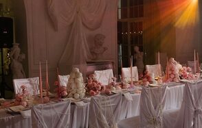 Арт-Плейс Фактура. Просторный лофт в классическом стиле. Столовая зона с длинным столом, накрытым белой скатертью и украшенным розовыми цветами и свечами. На заднем плане скульптуры и драпировки. На фоне виден мягкий свет, создающий атмосферу. Стулья обтянуты белыми чехлами.