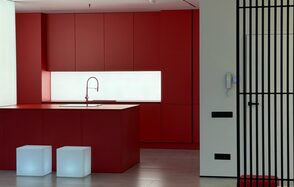 Арт-Плейс Фактура. Современный лофт с видом на весь город. Красная кухня с островом, встроенная раковина и высокий смеситель. Шкафы с закрытыми фасадами. Два белых кубических стула. Перегородка из черных вертикальных прутьев справа. Белая стена со встроенным домофоном и черными розетками. Светлые полы.